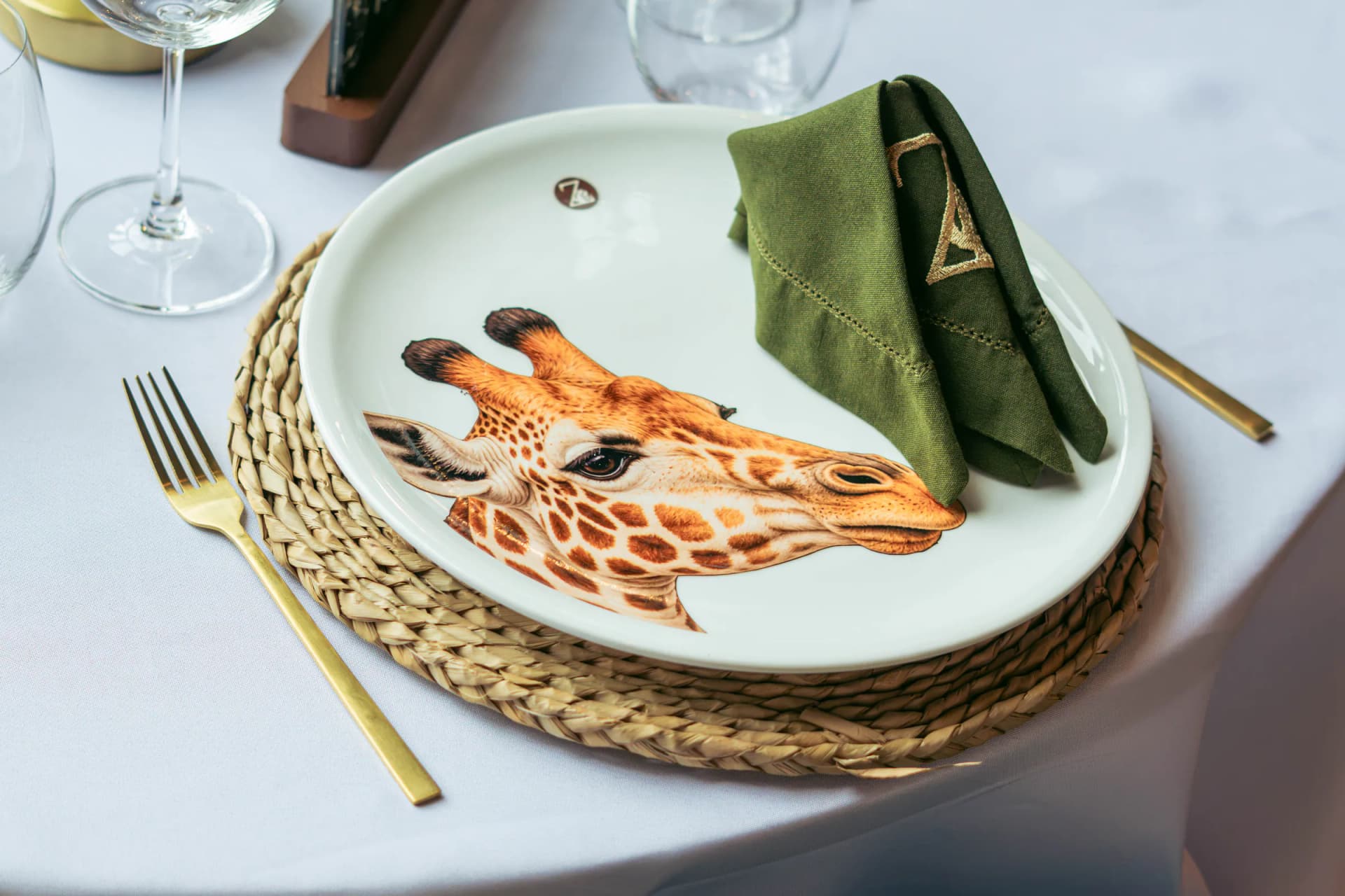 Plato de porcelana con la ilustración de una jirafa, servilleta verde con el isotipo bordado y cubiertos dorados