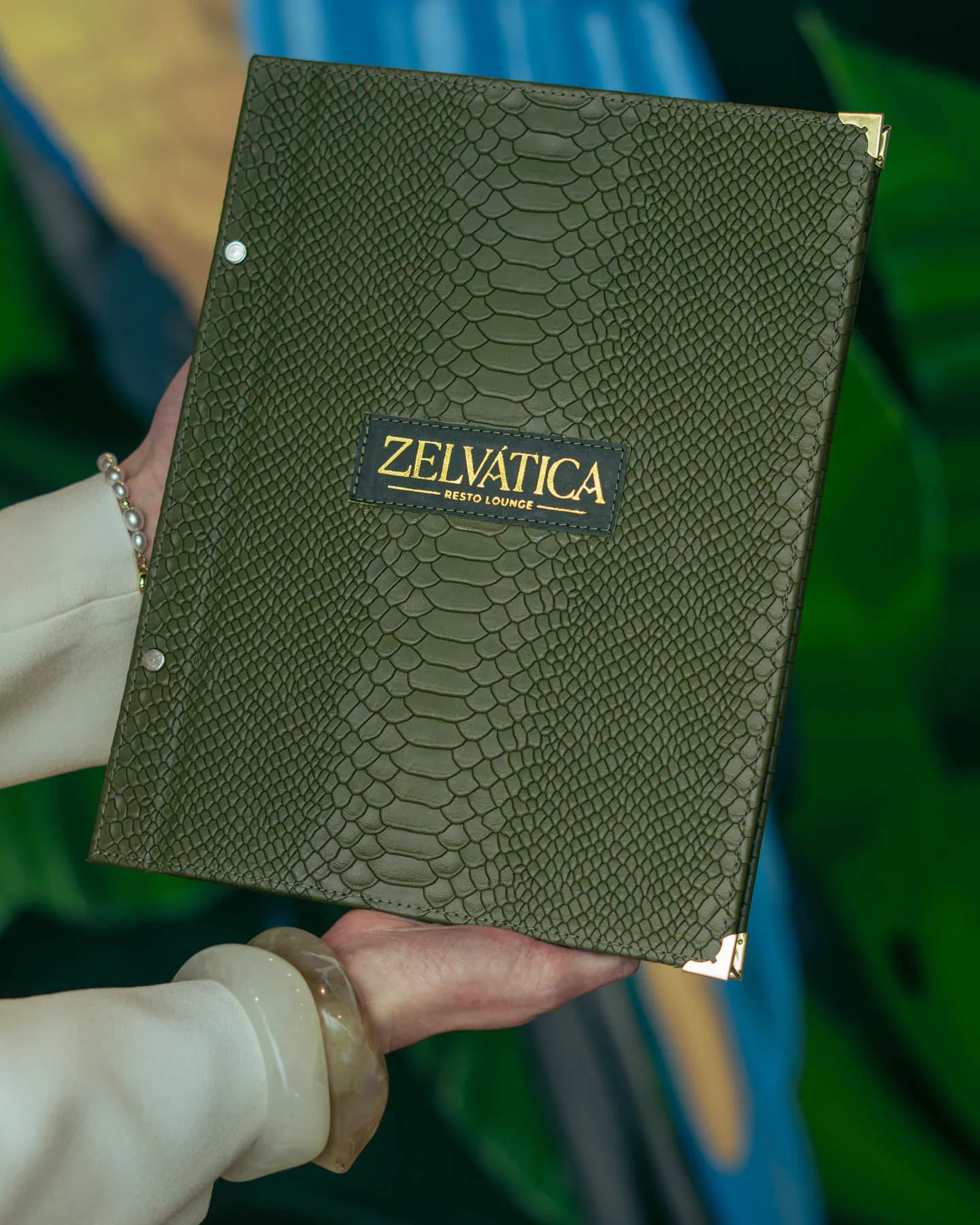 La carta de cuero verde con el logotipo dorado sostenida por una persona frente al mural