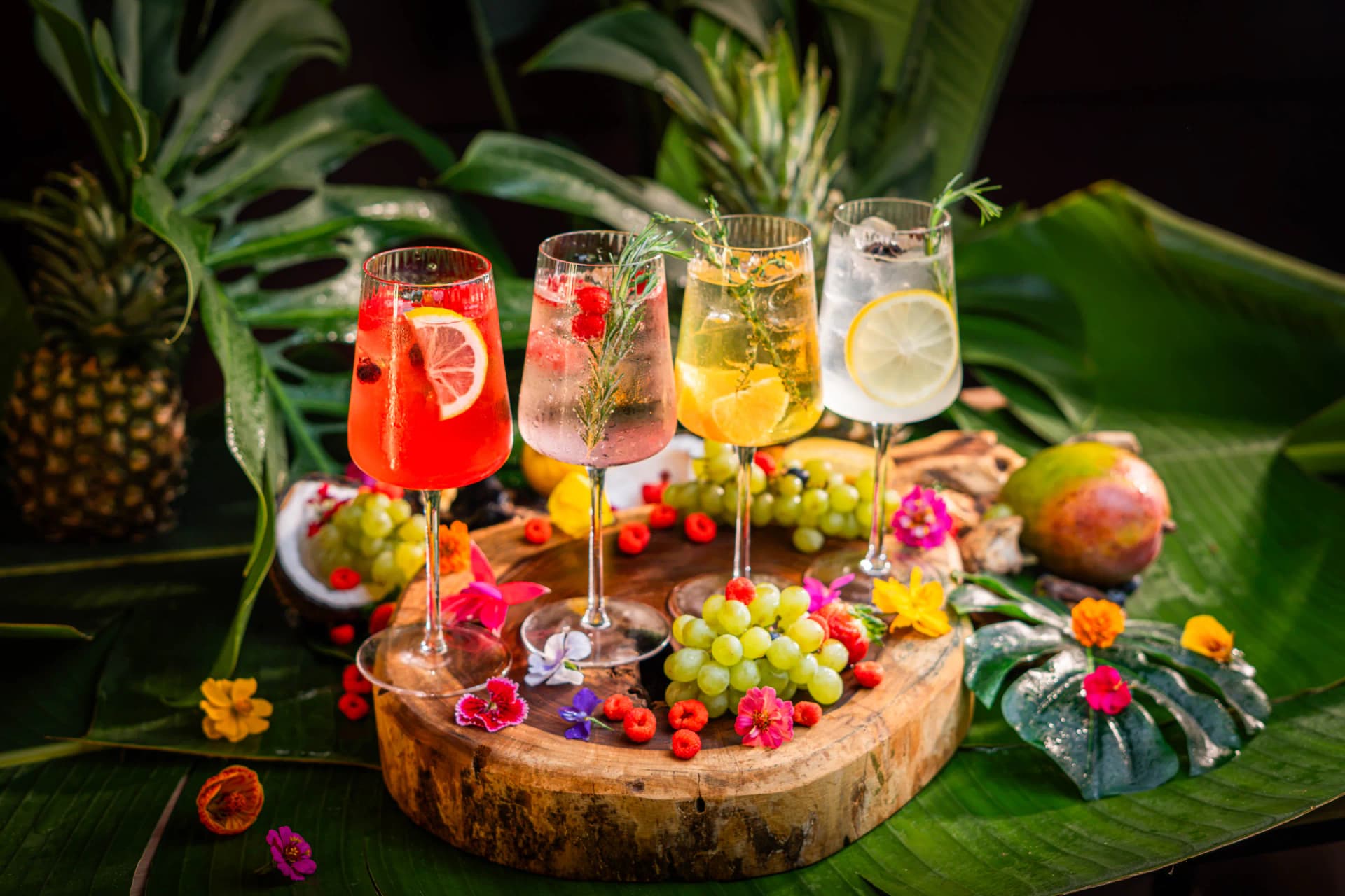 Cuatro copas de gin tonic de colores sobre un tronco cubierto de flores comestibles y frutas