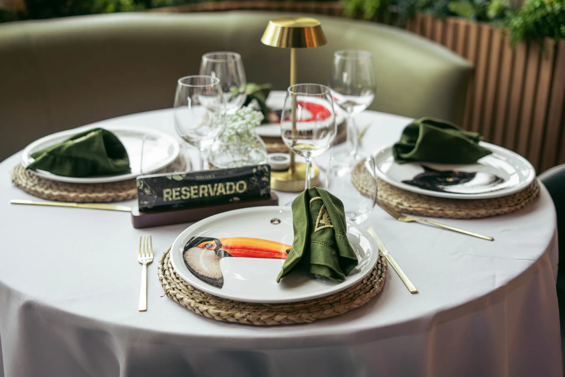 Mesa con un cartel de reservado, un plato ilustrado con un tucán, una lámpara de latón y un sofá verde oliva