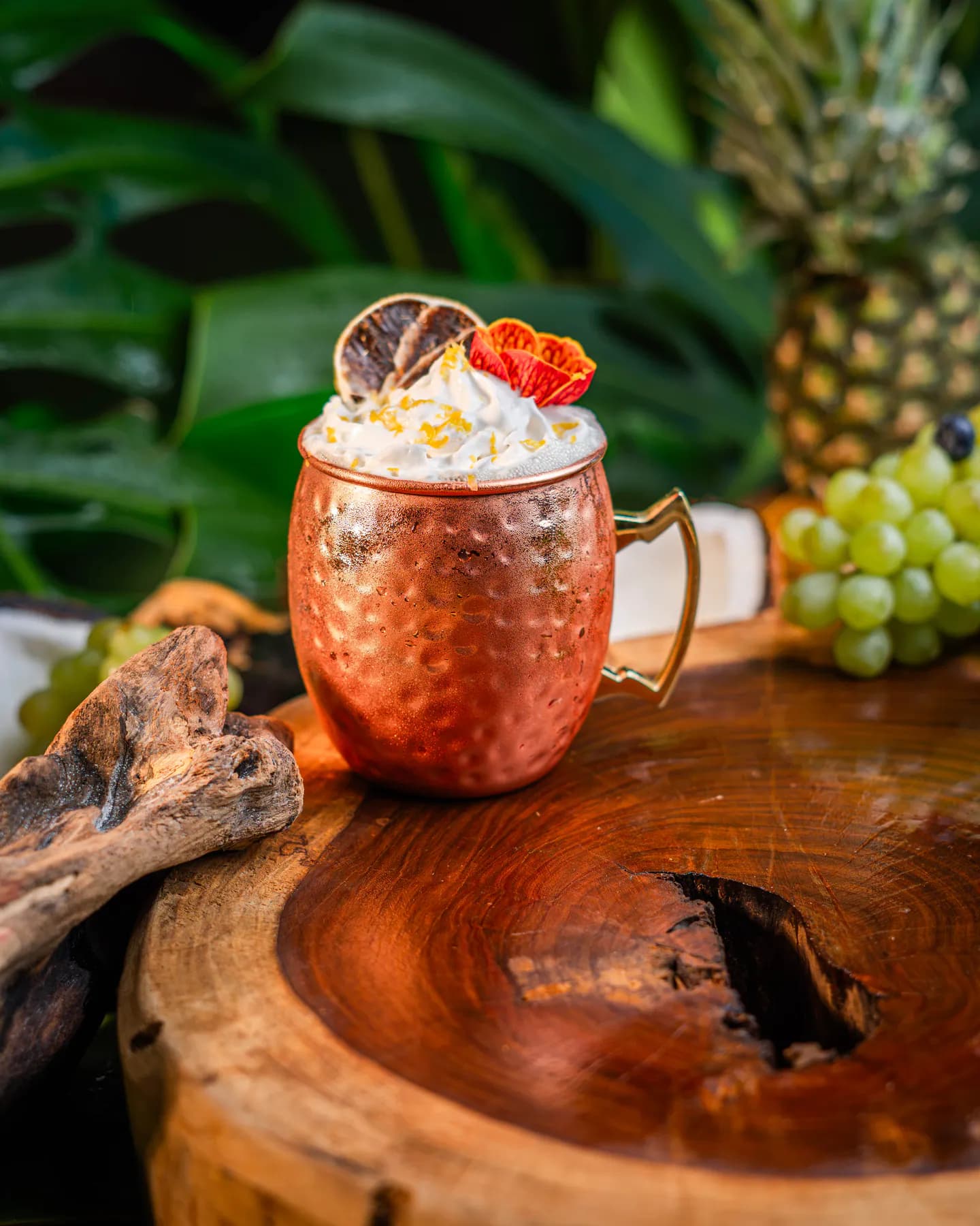 Trago servido en jarra de cobre martillado con crema y una flor, sobre un tronco