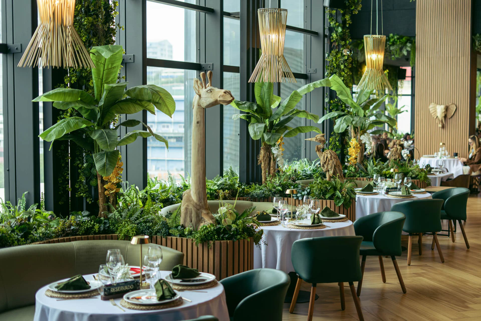 Vista general del salón con la jirafa de madera, lámparas de mimbre encendidas, vegetación y mesas servidas