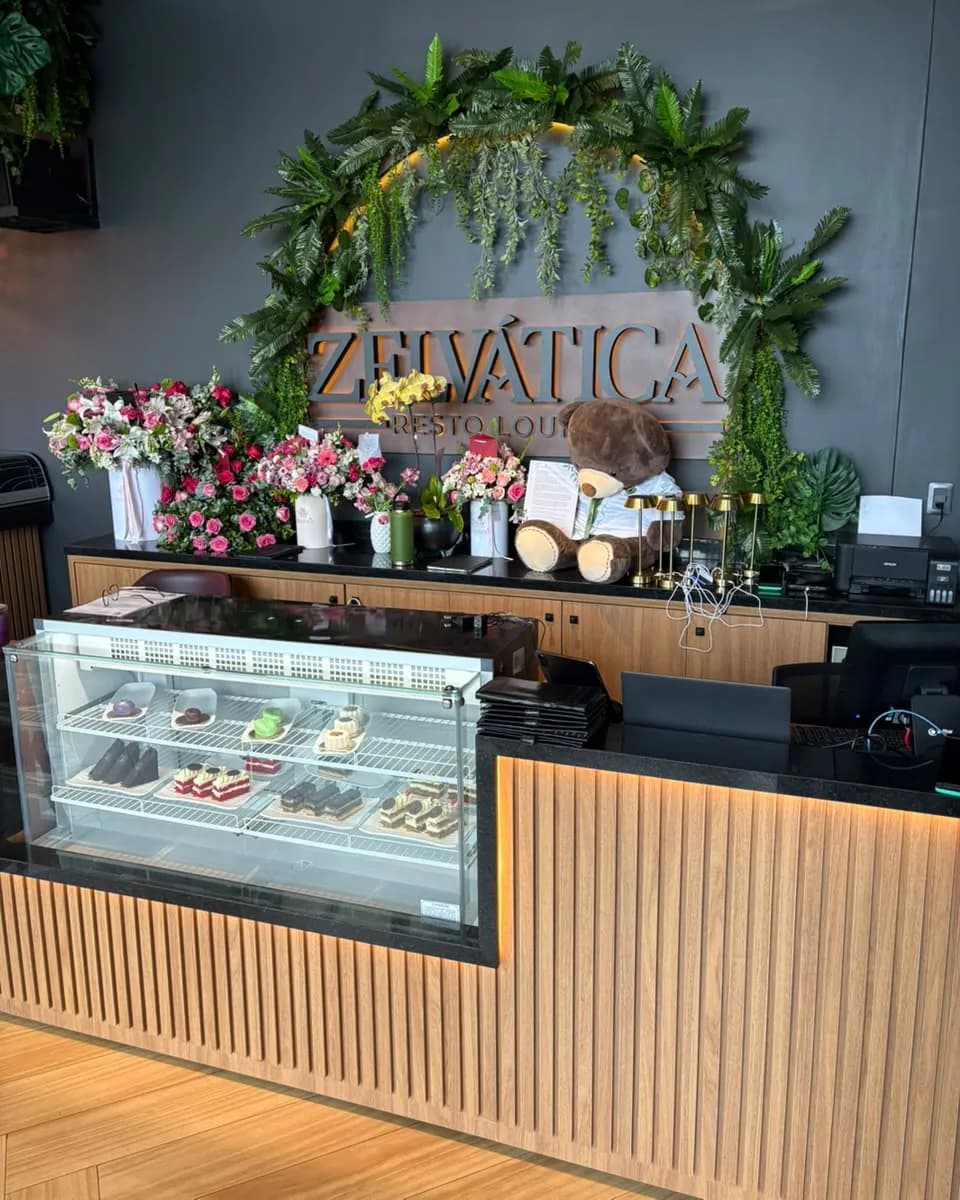 Mostrador de pastelería con la vitrina de tortas, ramos de flores y el cartel de Zelvática bajo un arco de follaje